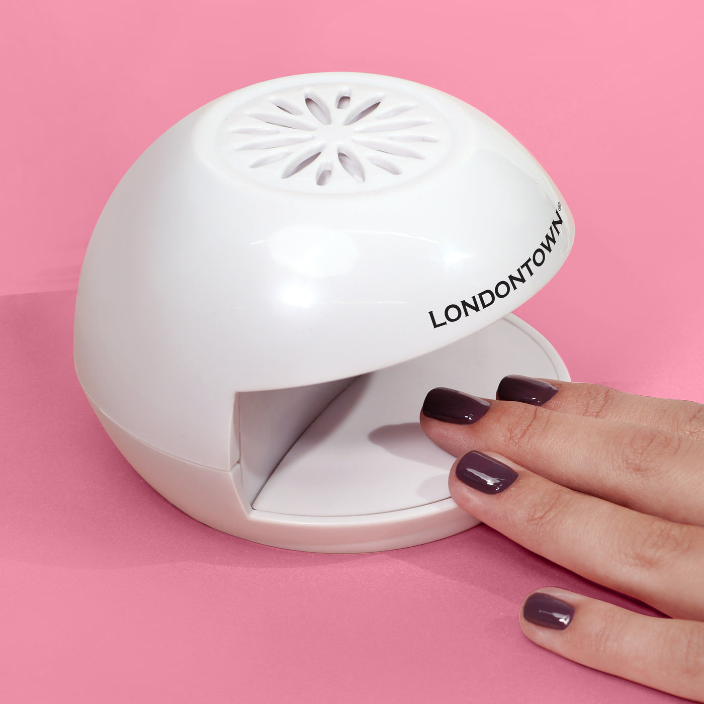 A hand with nails painted dark purple is placed under the "LONDONTOWN Flash Dry Nail Fan," a white semi-circular nail drying device, on a pink background—perfect for achieving salon-quality manis at home.
