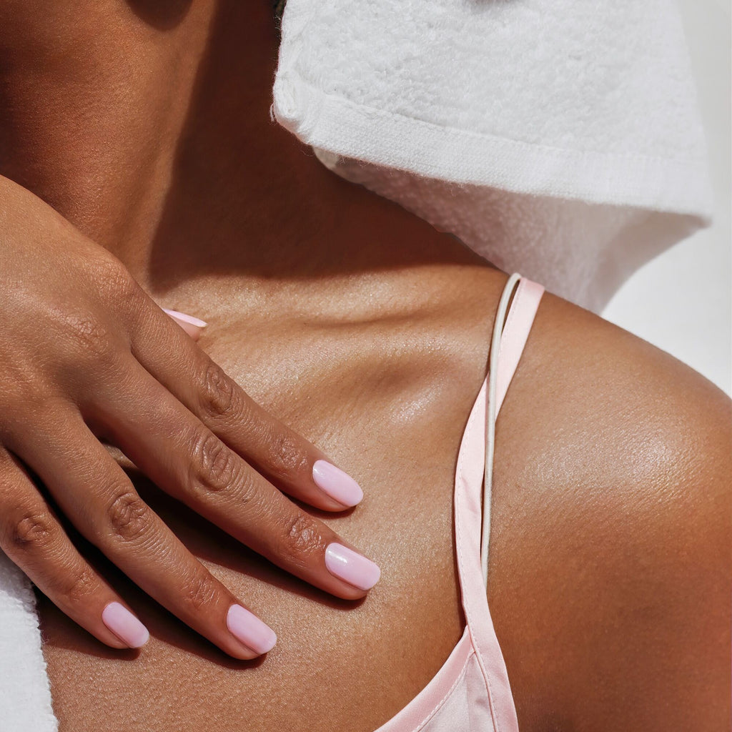 A person with light pink nail polish rests their hand on their upper chest, wearing a pink satin strap and a white towel over their shoulder.